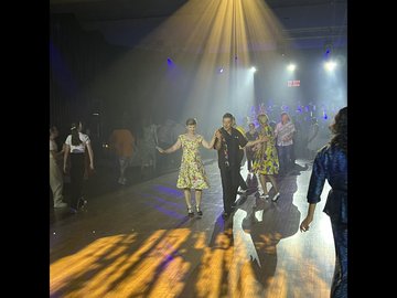 Couple in vintage attire dancing under a spotlight at a swing dance event, with other couples and attendees in the background, a stage light beam and light from overhead is illuminating the dance floor.