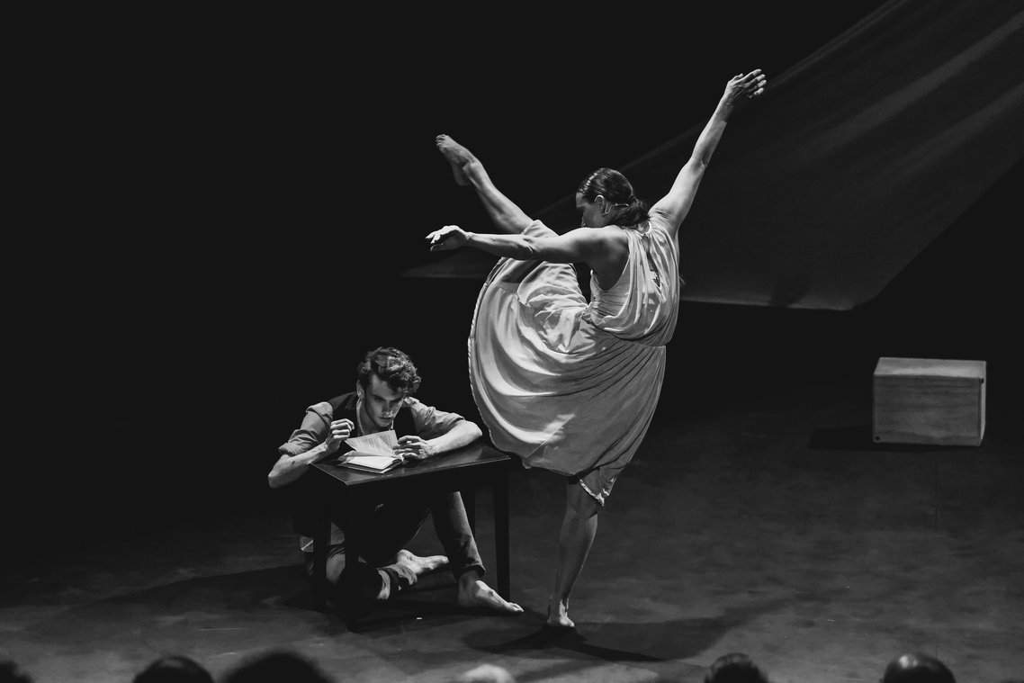 Ballet dancer mid-performance with man reading at desk, black and white stage photo.