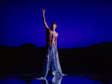 A shirtless male dancer in loose, patterned trousers, arms raised, poised mid-performance on a blue stage, a dark, mountainous backdrop behind him.
