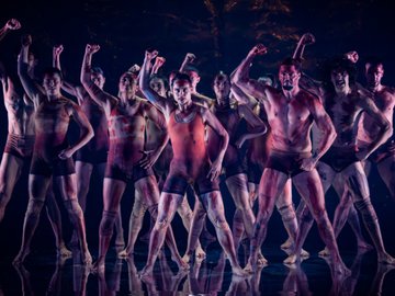 Dancers in a stage performance, wearing shorts and body paint, with raised arms and a dramatic expression. Some are shirtless and others wear red sports bras or tops.