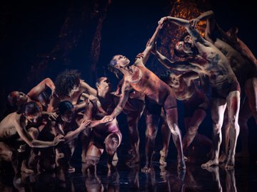 A group of dancers, painted in red and brown hues, perform a modern dance routine on a dimly lit stage, with fluid and expressive movements.