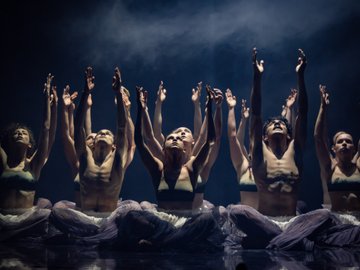 A dance performance featuring a group of cross legged performers with their arms raised upwards towards a dim, smoky background. They are wearing loose white pants. Some are shirtless and others wear sports bras.