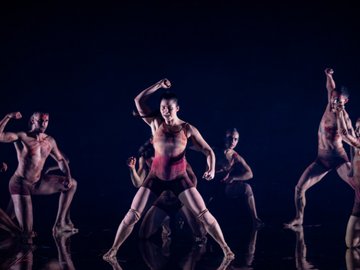 A modern dance performance with a group of diverse dancers, painted with red streaks, posing in dynamic and expressive poses on a dark reflective stage.