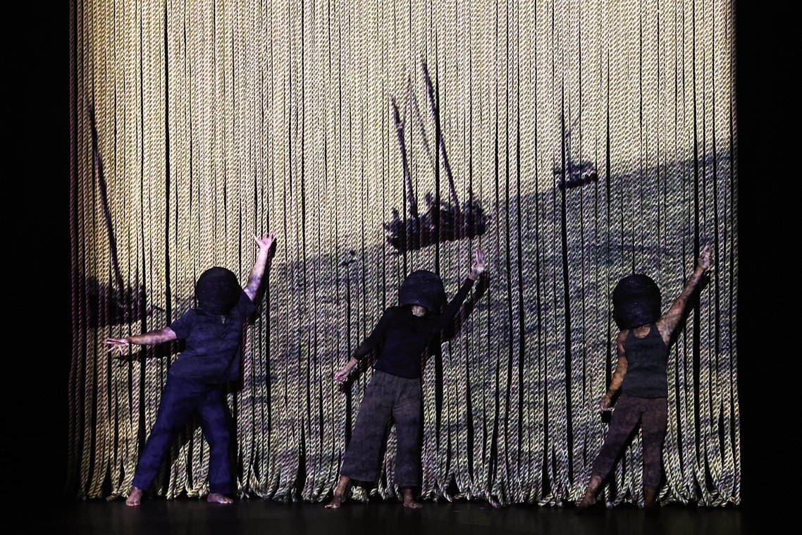 Three performers with black hoods obscuring their faces stand on stage behind a curtain of thick, light-colored ropes. A projection on the curtain shows two sailboats on a dark, choppy sea.