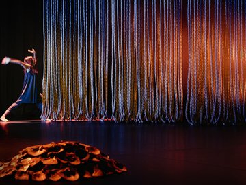 A dancer in a dark blue dress and pants performs on a stage in front of a curtain of many thick ropes, lit with blue and orange lights, with a sculptural element in the foreground.