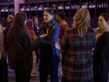 Group of women chatting at an indoor event, shelves of bottled beverages in background.