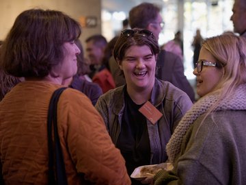 Three people smiling and talking at an indoor event; one holds a plate of food.