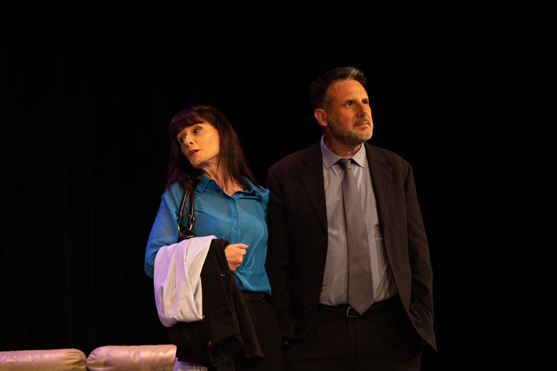A middle-aged man and woman in formal clothes. She has her hand around his back.