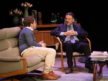 Two men sitting on couches in a living room. One man looks uncomfortable and has his hands clasped.