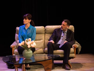 A middle-aged man and woman sitting on a leather couch with a vase of flowers in front. Neither of them look very happy.