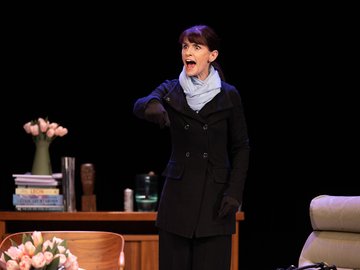 A middle-aged woman standing in a living room with a coat and scarf on. She has an angry look on her face.