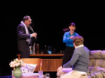 Two men and a woman in a living room. One is sitting with his back to camera. The other two are standing and look to be fighting.