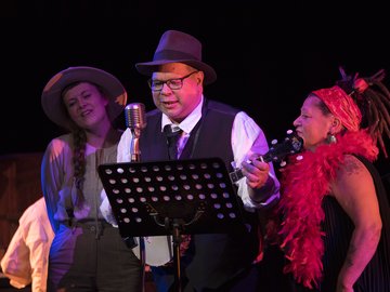 A trio of performers on stage, a woman with braided hair and a hat sings into a vintage microphone, a man in a vest and fedora plays a ukulele, and another woman with dreadlocks and a red feather boa sings into a microphone.