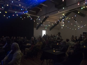 An audience sits at tables in a dimly lit room adorned with colorful string lights, watching a performance on a stage with a black backdrop.