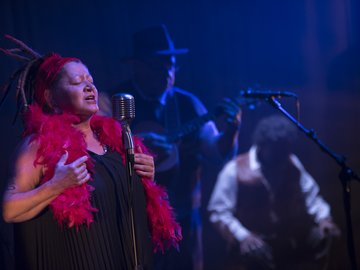 A singer with dreadlocks and a red feather boa stands before a vintage microphone, singing with eyes closed. In the blurry background, musicians play instruments under blue stage lights.