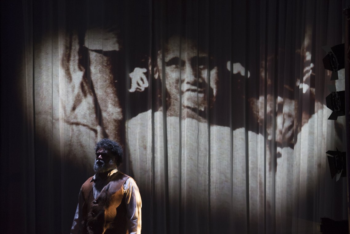 A man with a beard and curly hair stands on stage, bathed in light. Behind him, a large projection of a sepia-toned portrait of a person's face is displayed on a curtain.
