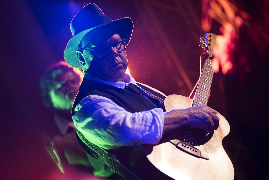A musician wearing a hat and glasses plays an acoustic guitar on stage, illuminated by coloured stage lights.