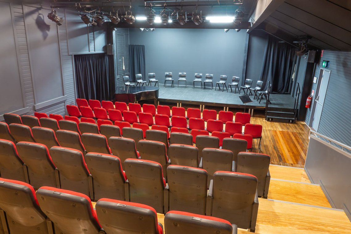 Interior view of a small theater with red and gray seats facing a stage with black curtains and a row of chairs, overhead lights and wooden floors.