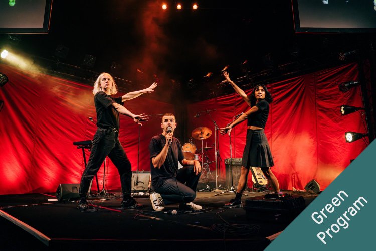Three performers on stage under red stage lighting: two with arms outstretched, flanking a third performer kneeling and holding a microphone, with a drum kit and musical equipment visible in the background.