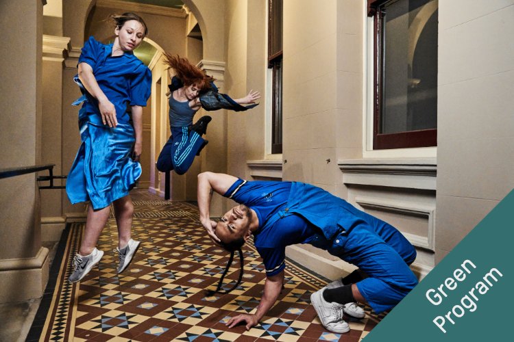 Three dancers in athletic poses within a historic architectural hallway with tiled floors, one dressed in a blue silk dress floating mid-air, a second dancer in a blue Adidas tracksuit leaping backwards, and a third in a blue jumpsuit arched backwards nea