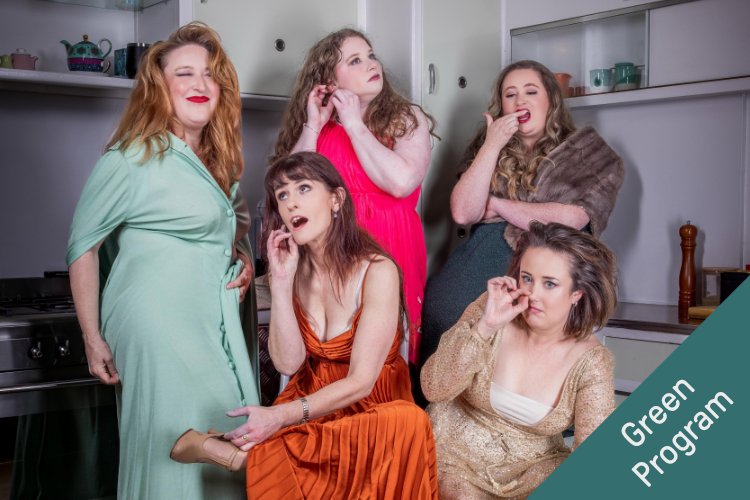 Five women in vintage-style clothing in a kitchen pose playfully, some touching their ears, mouths, and noses; the woman on the left wears a mint green button-down dress and laughs. The background includes kitchen cabinetry and appliances. A diagonal gree