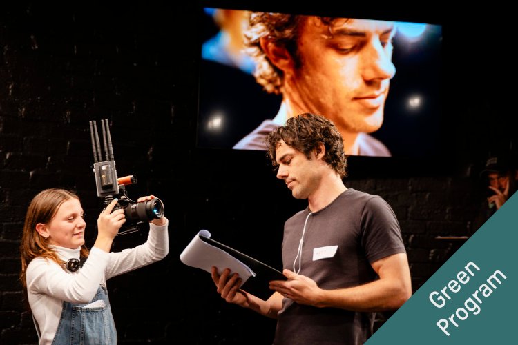 A young girl holds a professional camera with external antenna, smiling, focused on filming. A man beside her, holding a script and looking down, wears a lavalier microphone. Behind them, a screen displays a close-up of a man's face, with a dark brick wal