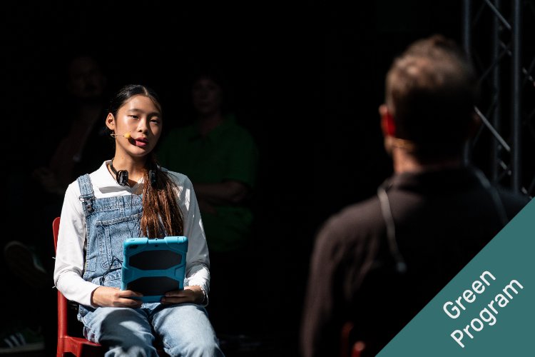 A young person wearing denim overalls over a white shirt sits in a chair, holding a tablet, with headphones around their neck. They are lit by a spotlight against a dark background. In the foreground, the back of another person's head is out of focus.