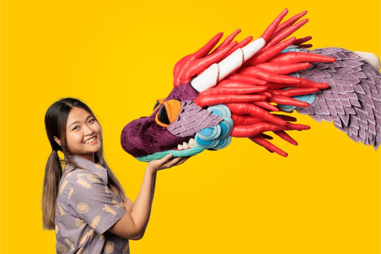 A young woman with dark hair smiling at the camera, with her hand on a colourful dragon puppet.