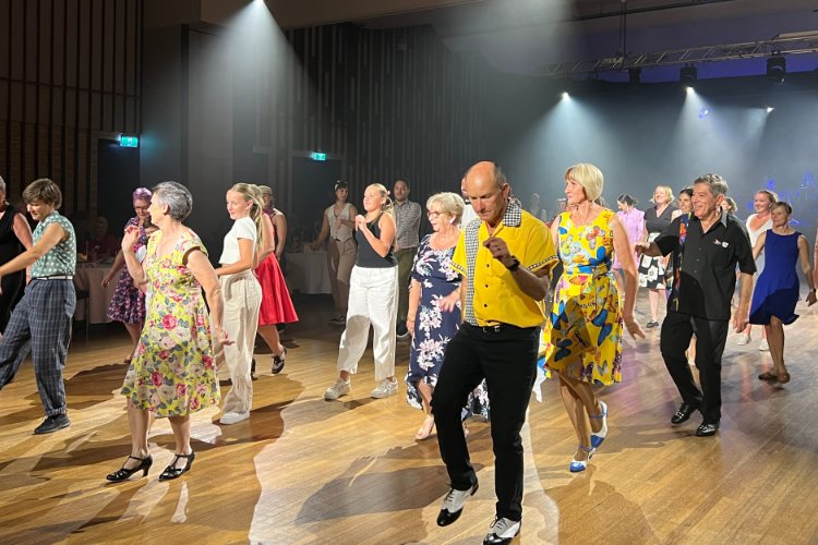 A diverse group of people dancing a swing dance in a brightly lit ballroom, with a man in a yellow shirt and black slacks as a prominent figure at the center of the frame.
