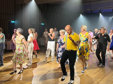 A diverse group of people dancing a swing dance in a brightly lit ballroom, with a man in a yellow shirt and black slacks as a prominent figure at the center of the frame.
