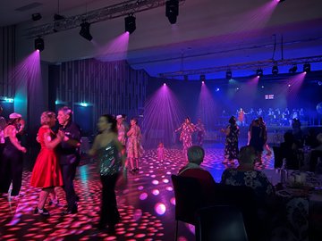 People dancing at a ballroom event with a live band on stage. The room is lit with purple and blue stage lights and a disco ball effect on the floor. Dancers are in couples and groups, enjoying the music. Audience members are seated at tables around.