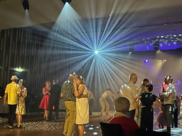 People dancing at a formal event under bright spotlights, some couples are embracing and others are talking.