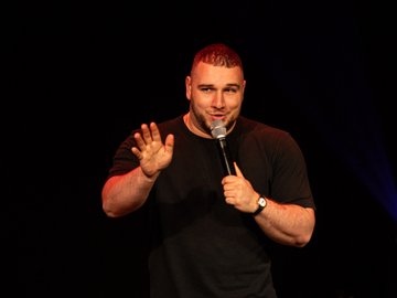 A bald man wearing a black t-shirt is performing stand-up comedy on a dark stage, holding a microphone in one hand and gesturing with the other. He is wearing a watch and illuminated by stage lighting.