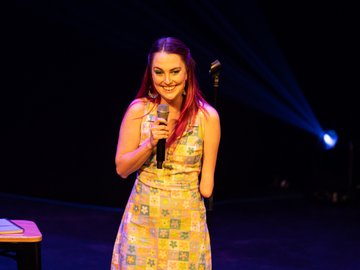 A woman with pink hair and a yellow floral dress stands on a stage holding a microphone, smiling, with a stage light in the background. She has a limb difference on her left arm.