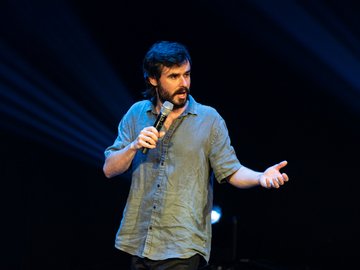 A man with a beard and dark hair wearing a light green shirt, holding a microphone and gesturing with his right hand on a dark stage with stage lights.
