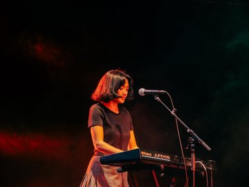 A woman with mid-length dark hair plays a keyboard standing up. A microphone stands nearby.
