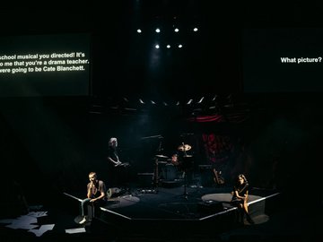 Rock band instruments on a stage with dim lighting. One person stands at keyboard and two people sit on the edge of the stage with microphones.