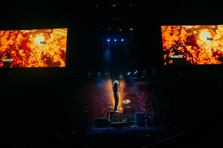 Rock band instruments on a stage with dim lighting. A man with a wolf mask stands on top of the bass drum with a microphone in his hand.