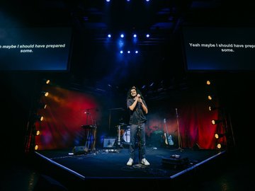 Rock band instruments on a stage with theatrical lighting. A person stands at the front of the stage with a microphone. Two screens above display the text 'Yeah maybe I should have prepared some'.