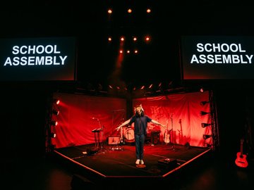 Rock band instruments on a stage with red lighting. A person with a wolf mask stands at the front with their arms out to the side. Two screens with the words 