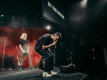 A band playing on a stage. The lead singer bends over singing into the microphone. A screen above displays 'By the rocks'