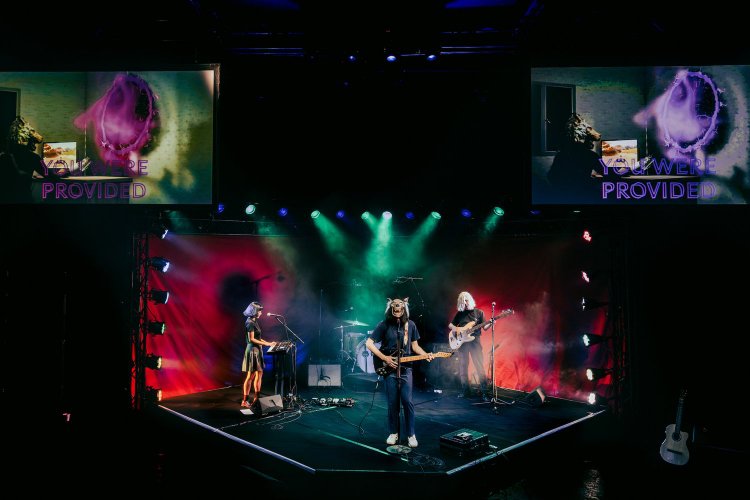 A 3-piece rock band on stage with theatrical lighting. The lead singer wears a wolf mask. Two screens appear above with the text 'You were provided'.