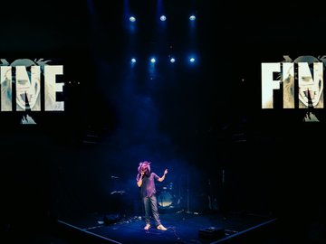 Rock band instruments on a stage with dim lighting. A person with a wolf mask stands with a microphone. Two screens appear above with the text 'Fine'.
