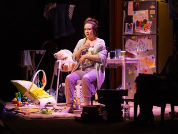 A woman dressed in casual pajamas, looking pensive, holds a baby doll on a stage set designed to resemble a cluttered living room with baby items and a refrigerator decorated with children's artwork.