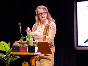 A woman with glasses and light brown hair stands at a wooden podium speaking into a microphone. She wears a beige blazer with a striped shawl, a white shirt, and tan pants. On the podium are several potted cacti.