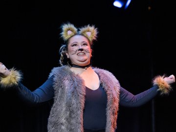 A person in a cat costume, wearing ear headbands, cat whiskers drawn on their face, faux fur vest and wristbands, arms outstretched, on a stage with a dark background.