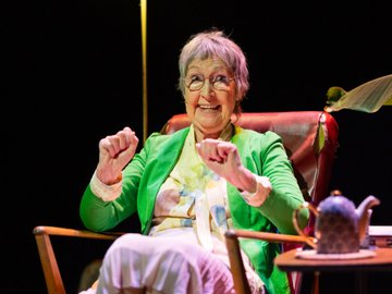 An elderly woman with short gray hair and glasses, wearing a green jacket, sits in a wooden armchair with a red leather back and a table with a teapot in front of her, raising her fists and smiling widely.