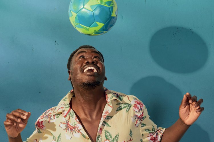 A Sudanese man in a floral shirt looks up at a soccer ball in the air, his arms outstretched with palms facing up, against a turquoise background.