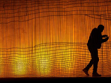 Silhouette of a person against a bright orange backdrop with vertical lines and a grid-like net overlay, person is holding something in their hands.