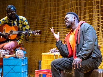 A South Sudanese man in a black jacket and orange shirt, gesturing as he sits on luggage, with another man playing a guitar in the background, in front of a yellow net.